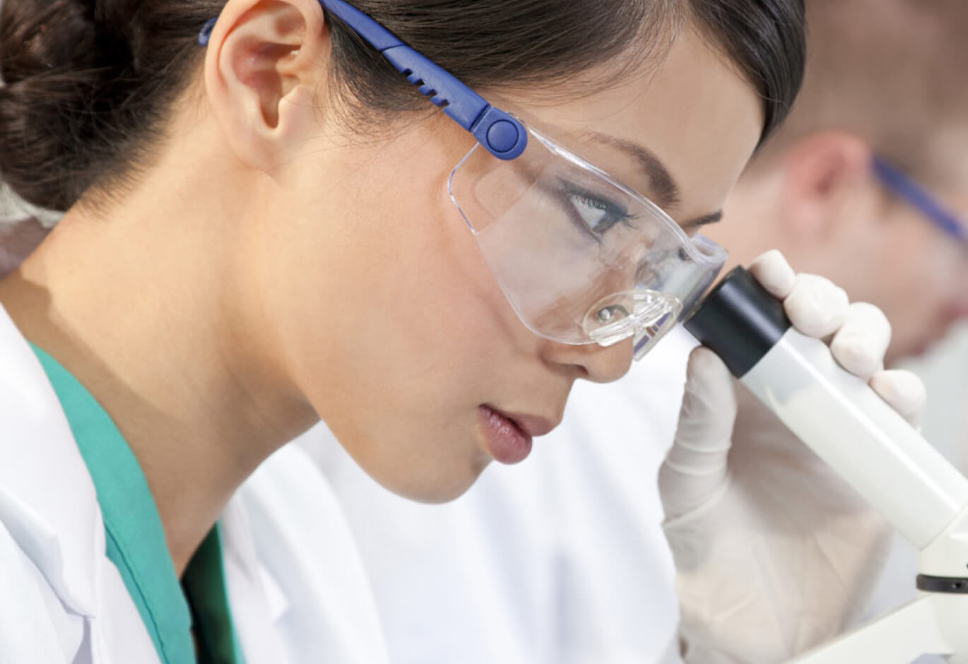 A woman wearing safety googles looking into a microscope.
